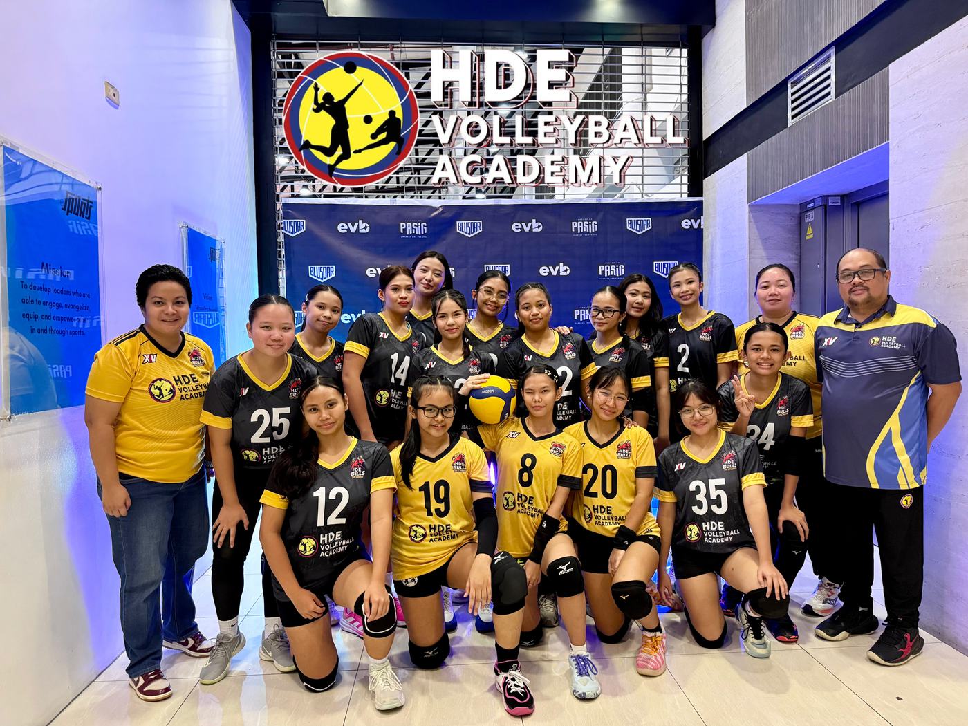 An HDE team in black and gold with their coaches under the HDE Volleyball Academy sign