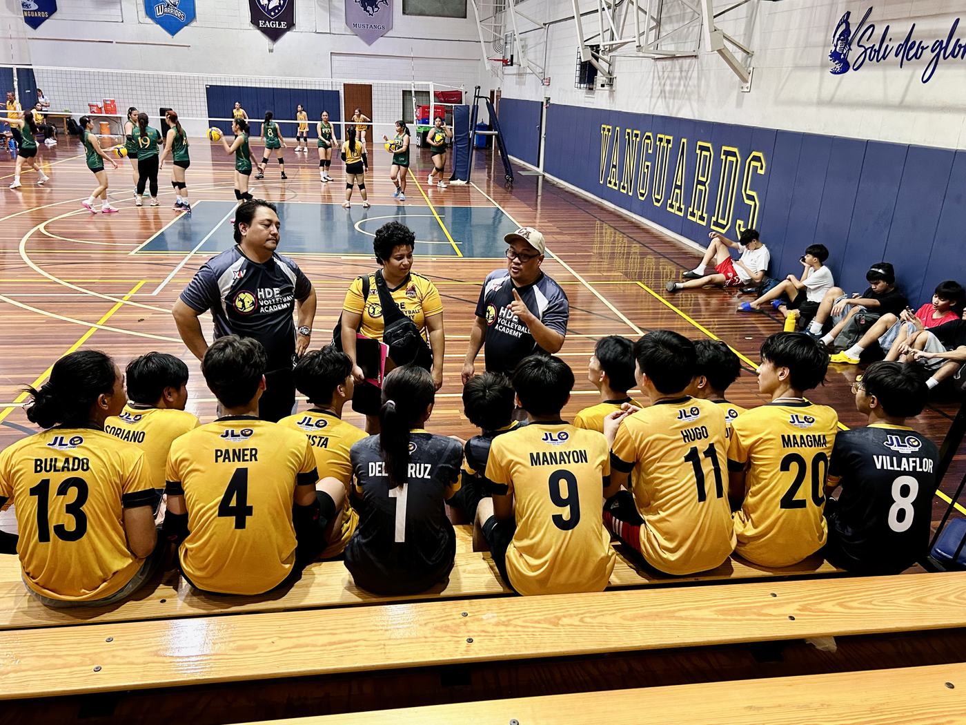 HDE players in gold seated courtside in front of their coaches