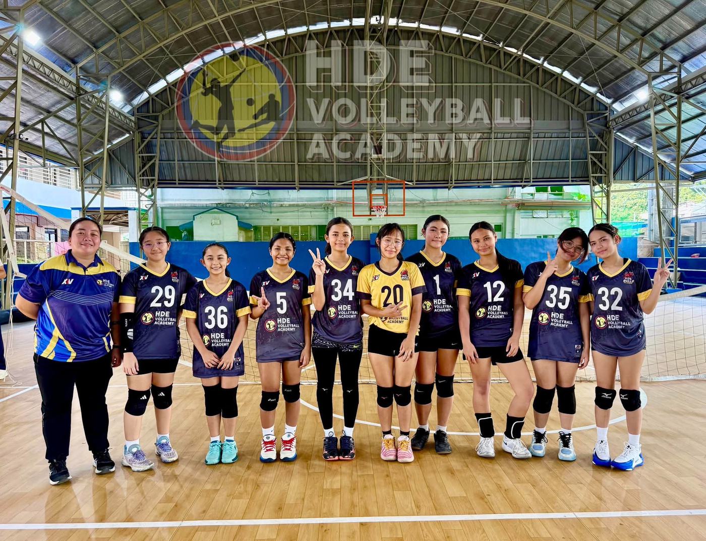 An HDE team in navy and black jerseys with their coach under the HDE Volleyball Academy sign