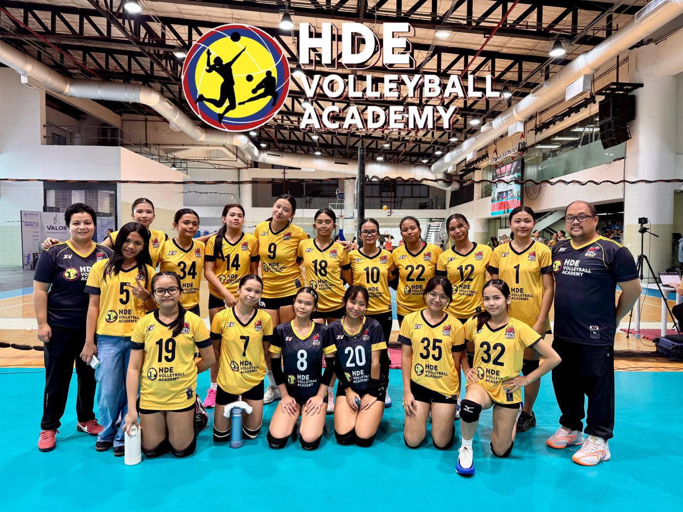An HDE girls team in yellow and black kneeling with their coaches on a teal court
