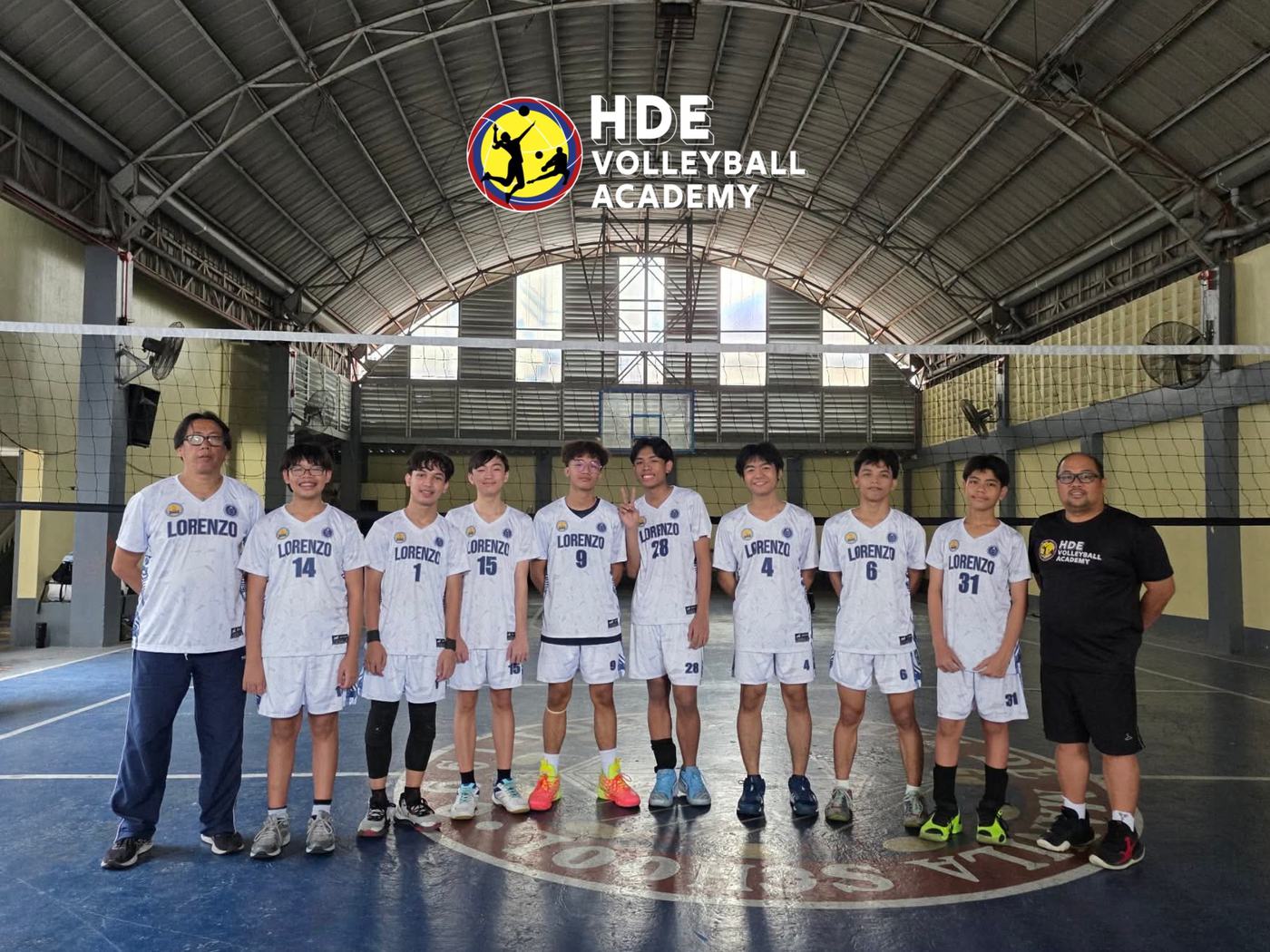 A school volleyball team in white uniforms with HDE coaches under the HDE Volleyball Academy sign