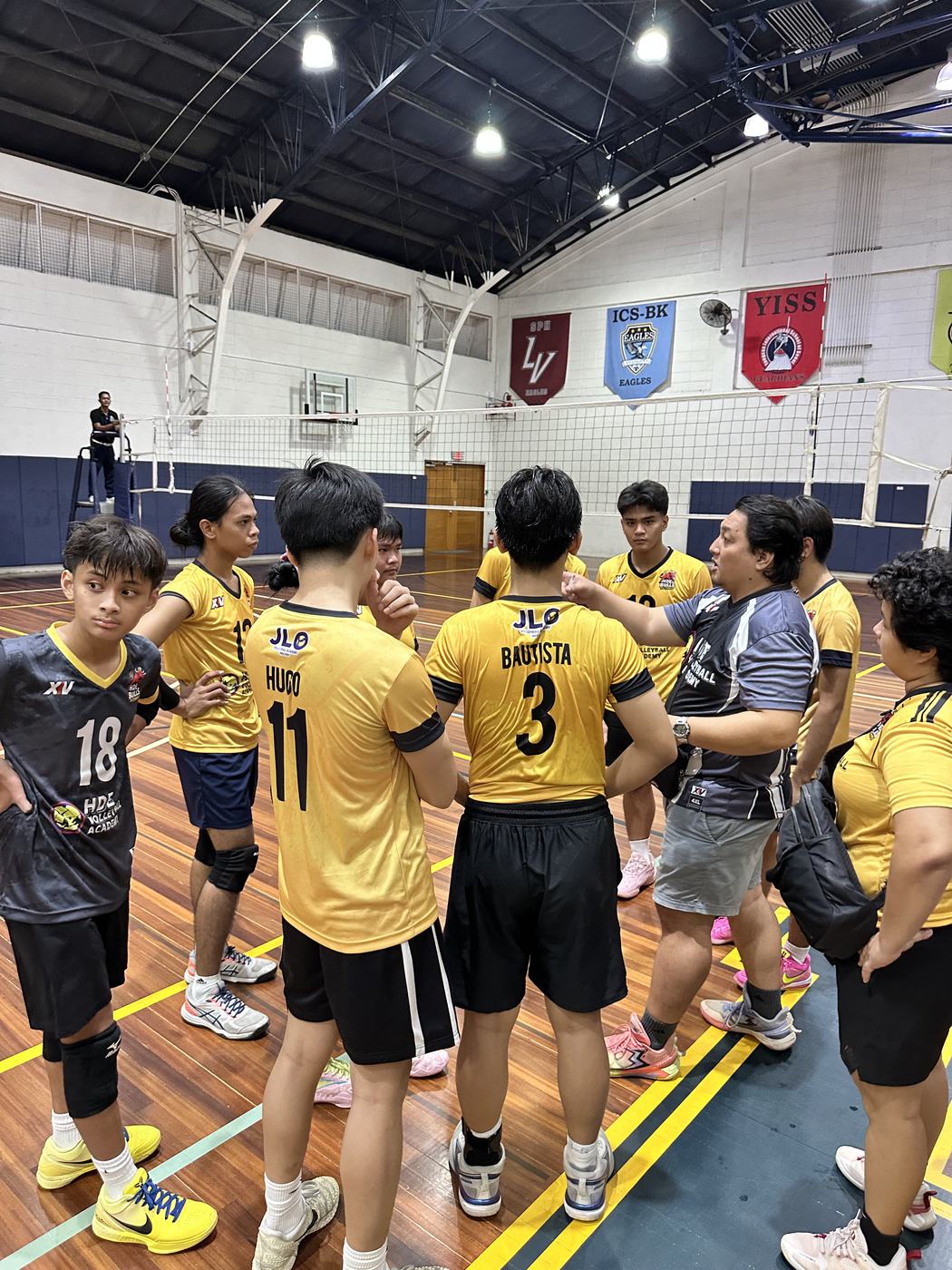 An HDE team in black and gold jerseys gathered with their coach in a gym