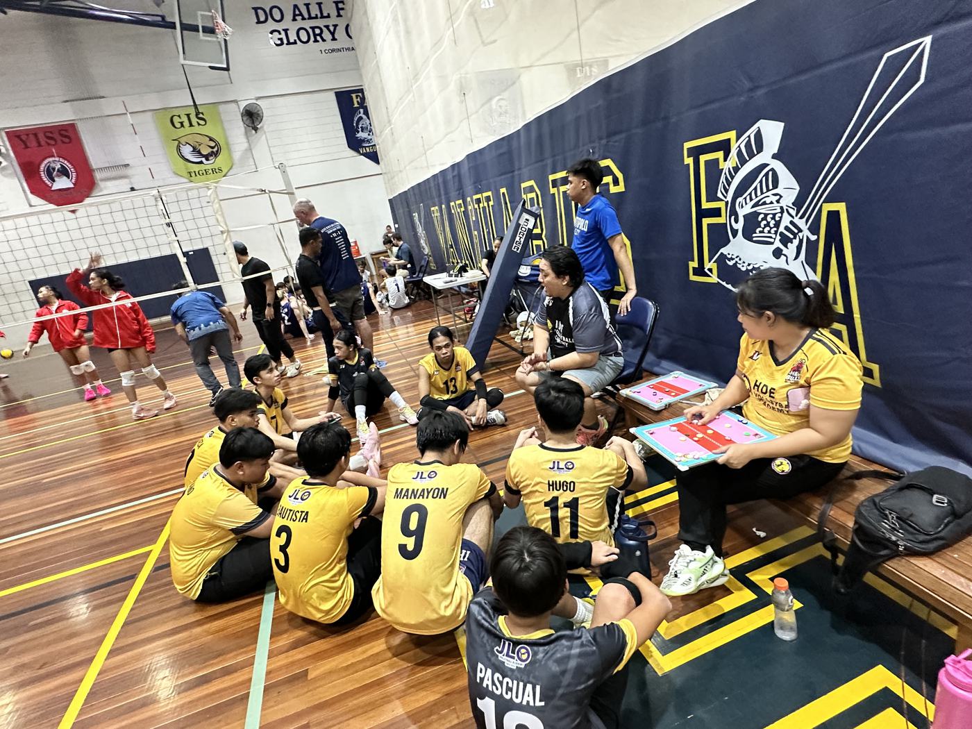 An HDE coach with a clipboard talking to seated players courtside during a tournament